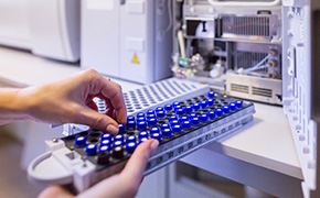 Reference Materials A close-up image of a person's hand inserting a blue and black sample tray into a laboratory instrument. The background shows part of the machine with visible components, emphasizing a laboratory setting.