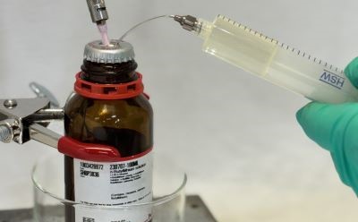 Laboratory extraction of an organolithium reagent A close-up image of a laboratory setting showing a brown glass bottle of organometallic reagents with a white label and a Sure/Seal™ cap. A syringe is being used to extract liquid from the bottle, with a thin needle supplying inert gas inserted through the cap. The person handling the syringe is wearing a green glove, indicating safety precautions. The background is blurred, focusing on the bottle and syringe, with a glass container visible beneath the bottle.
