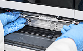 HPLC Columns HPLC column is installed in lab instrument. The hands of a lab worker are shown wearing blue nitrile gloves.