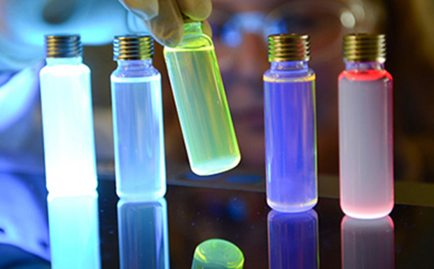 Vials of OLED materials lit up by UV light A close-up image of several glass vials with metallic caps, illuminated by colorful lights. The vials contain liquids that glow in shades of blue, purple, and red, creating a visually striking effect against a reflective surface. A gloved hand is positioned near the vials, suggesting a scientific or experimental context.