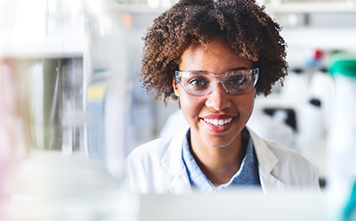 mRNA-analytics-and-quality-control A person wearing a lab coat and safety glasses is working in a laboratory setting, surrounded by scientific equipment and glassware. The background features blurred laboratory elements, emphasizing a professional research environment.