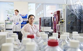 分析開発サービス 白色と赤色のキャップの付いた多数のボトルが置いてあるバイオ医薬品調製・サンプル分析のための分析開発ラボで働いている1名の男性科学者と1名の女性科学者。